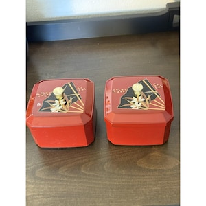 May include: Two red, octagonal boxes with lids. Each lid features a black and gold design with a mountain, fan, and floral motif. The boxes have gold knobs and are likely for storage or display.