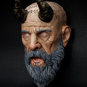 Puede incluir: Escultura detallada de un rostro envejecido con cuernos y barba gris. La cara está pintada en tonos marrones y beige. Los cuernos son de color dorado metálico y los ojos blancos. La escultura tiene un aspecto envejecido, con inscripciones en la cabeza.