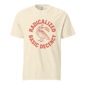 Puede incluir: Camiseta color crema con un gráfico circular rojo. El gráfico presenta un pájaro sosteniendo una ramita y las palabras "RADICALIZED BY BASIC DECENCY" en un arreglo circular. La camiseta tiene cuello redondo y mangas cortas.