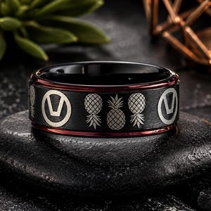 Puede incluir: Un anillo de banda negro con un borde interior rojo y un patrón grabado con láser que repite piñas y símbolos circulares con la letra V. El anillo tiene un acabado exterior negro mate y un borde interior rojo pulido.