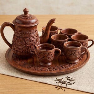 Könnte beinhalten: Ein geschnitztes Holz-Teeservice in Dunkelbraun, bestehend aus einer Teekanne, sechs Tassen und einem runden Tablett. Das Set weist Blumen- und Blattschnitzereien auf. Lose Teeblätter sind auf dem Tisch verstreut.