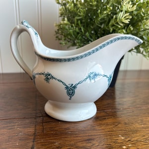 May include: White ceramic gravy boat with a curved handle and spout. The boat is decorated with teal floral and geometric patterns along the rim and body. It sits on a small, round base, and is a classic piece for serving sauces.