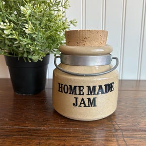 May include: A tan ceramic jar with a cork lid and a metal handle. The words "HOME MADE JAM" are printed in black on the front. A small potted plant is in the background.
