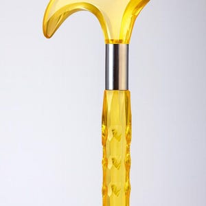 Puede incluir: Un bastón amarillo transparente con un mango curvo. El mango y el eje están facetados, lo que le da al bastón una apariencia de joya. Una banda de metal plateado conecta el mango con el eje. El bastón está sobre un fondo blanco.