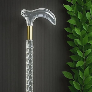 Puede incluir: Bastón transparente y curvado con una banda metálica dorada y un eje texturizado. El bastón se encuentra sobre un fondo oscuro con una planta verde y frondosa a la derecha.