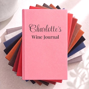 May include: A stack of rectangular journals with textured covers in various colors, including pink, red, orange, brown, blue, and white. The top pink journal features the black printed text Charlotte's Wine Journal in a mix of script and serif fonts.