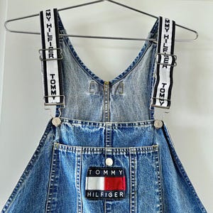 May include: A denim overall dress with adjustable straps featuring the Tommy Hilfiger logo in white text on a black and white striped background. The dress has a front pocket with the Tommy Hilfiger logo patch in red, white, and blue.