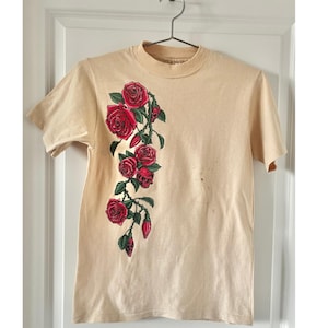 May include: A beige t-shirt with a crew neck and short sleeves. A detailed illustration of red roses and green stems adorns the left side of the shirt. The shirt is hanging on a metal hanger against a white door.