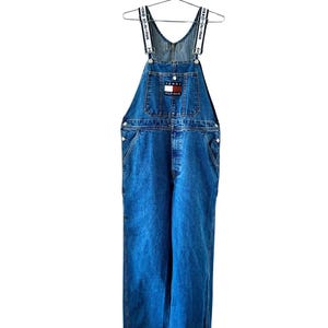 May include: Blue denim overalls with white and navy straps. The overalls feature a front pocket with a Tommy Hilfiger logo patch. The garment is hanging on a metal hanger against a white background.