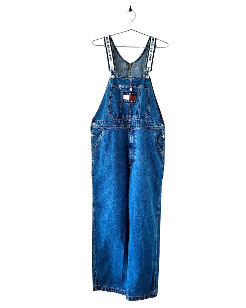 May include: Blue denim overalls with white and navy straps. The overalls feature a front pocket with a Tommy Hilfiger logo patch. The garment is hanging on a metal hanger against a white background.