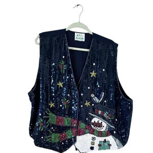 May include: A navy blue sequined vest with a Christmas snowman design. The vest features a snowman with a top hat and scarf, surrounded by stars and holly. The vest is hanging on a white hanger.