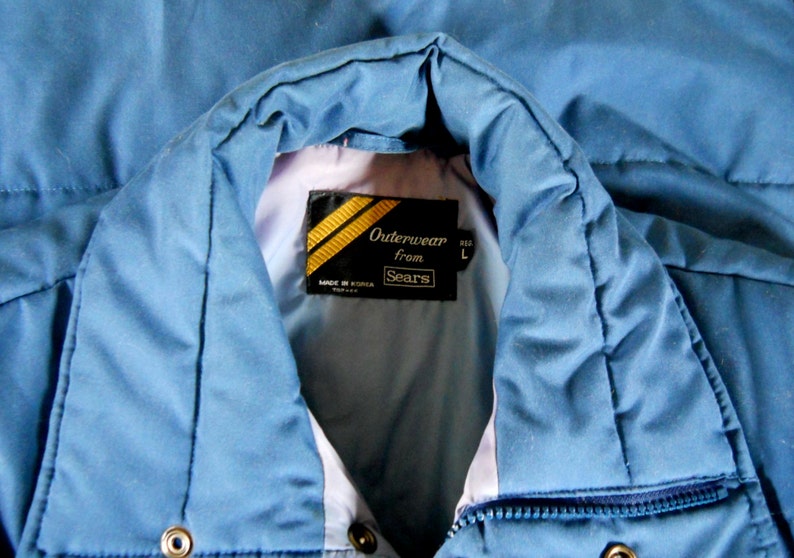 May include: A blue puffer jacket with a zipper closure and a label that reads "Outerwear from Sears" with gold stripes. The label also says "Made in Korea" and "Size L".