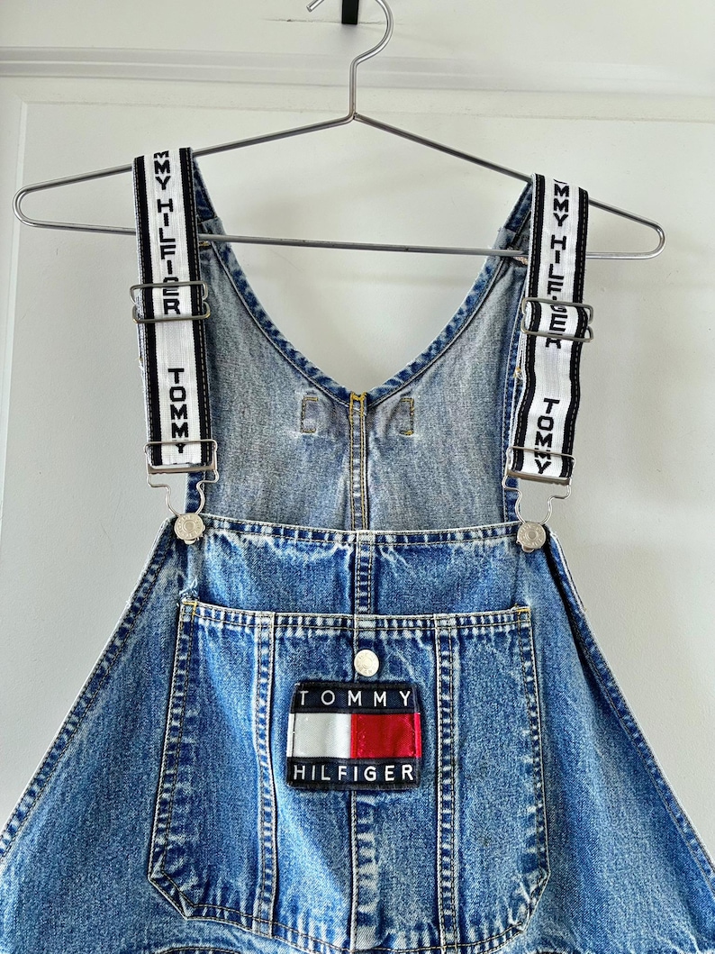 May include: A denim overall dress with adjustable straps featuring the Tommy Hilfiger logo in white text on a black and white striped background. The dress has a front pocket with the Tommy Hilfiger logo patch in red, white, and blue.