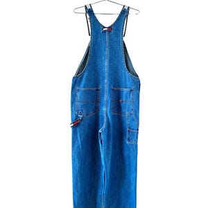 May include: Blue denim overalls with a classic design, featuring multiple pockets and adjustable straps. The overalls have a relaxed fit and are hanging on a metal hanger. The Tommy Hilfiger logo is visible on the front and side.