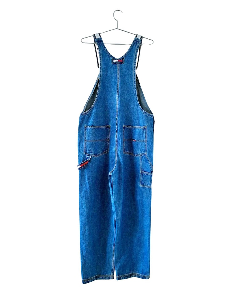 May include: Blue denim overalls with a classic design, featuring multiple pockets and adjustable straps. The overalls have a relaxed fit and are hanging on a metal hanger. The Tommy Hilfiger logo is visible on the front and side.