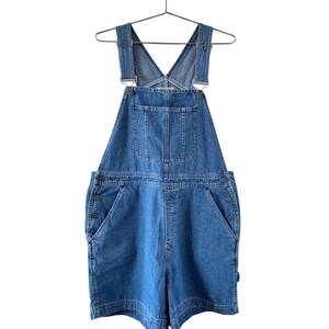 Puede incluir: Pantalones cortos de peto vaqueros azules con tirantes ajustables y m&uacute;ltiples bolsillos. Los pantalones cortos tienen un dise&ntilde;o cl&aacute;sico con cierres de bot&oacute;n en los laterales. La prenda est&aacute; colgada de una percha met&aacute;lica sobre un fondo blanco.