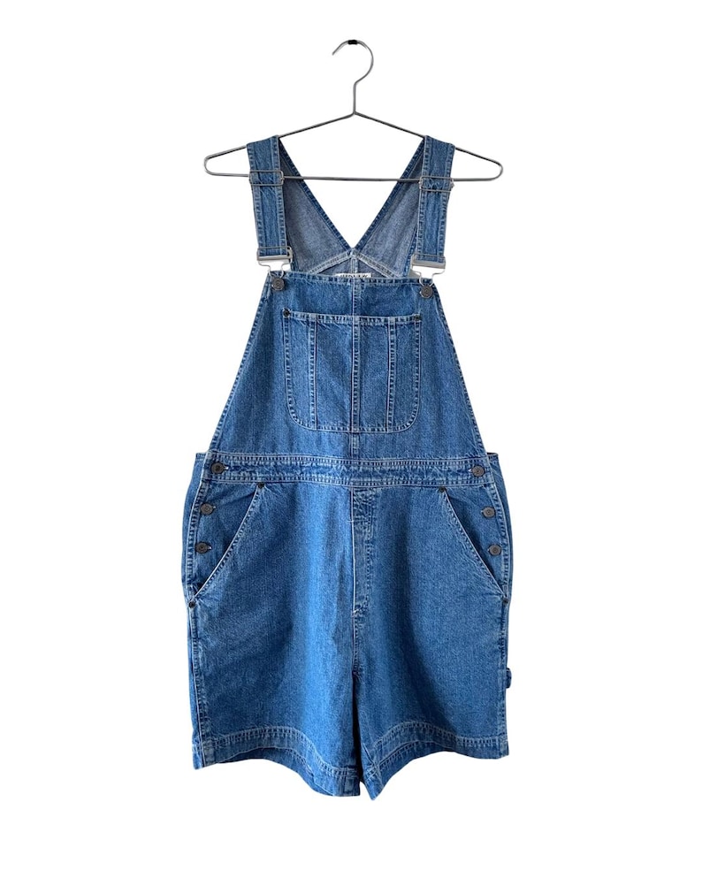 Puede incluir: Pantalones cortos de peto vaqueros azules con tirantes ajustables y m&uacute;ltiples bolsillos. Los pantalones cortos tienen un dise&ntilde;o cl&aacute;sico con cierres de bot&oacute;n en los laterales. La prenda est&aacute; colgada de una percha met&aacute;lica sobre un fondo blanco.