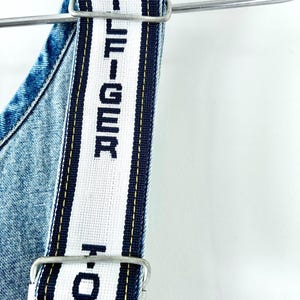 May include: Close-up of denim overalls with white straps featuring the text "HILFIGER" in black. The straps are trimmed with navy blue and yellow stripes. The overalls are hanging on a metal hanger against a white background.
