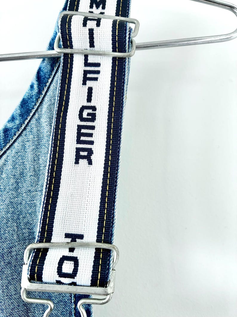 May include: Close-up of denim overalls with white straps featuring the text "HILFIGER" in black. The straps are trimmed with navy blue and yellow stripes. The overalls are hanging on a metal hanger against a white background.