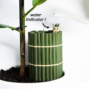 May include: A cylindrical green plant watering device with a bamboo-like texture and light-colored bands. It features a small, smiling character figure on the rim labeled water indicator! with an arrow pointing to the water being poured inside.