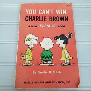 May include: Vintage paperback book titled "You Can't Win, Charlie Brown" with a red cover. The cover features a cartoon illustration of Charlie Brown and two other characters. The book is a "Peanuts" book by Charles M. Schulz.