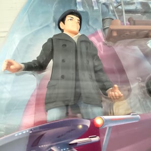 May include: A plastic action figure of a man wearing a black coat and a black cap. The figure is standing in front of a purple and silver spaceship. The figure is in a clear plastic package.