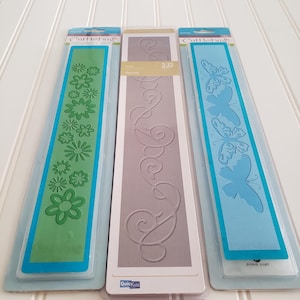 May include: Three long rectangular embossing folders in original packaging. The left folder is green with a flower pattern, the center folder is grey with a flourish design labeled 2.12, and the right folder is light blue with a butterfly pattern.