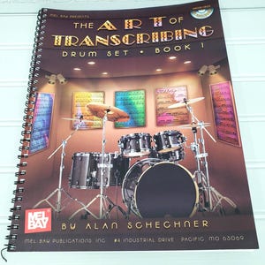 May include: A spiral-bound book titled "The Art of Transcribing Drum Set Book 1" featuring a drum set illustration. The cover has text in gold and brown tones, with a drum set image in the background. The book is from Mel Bay Publications.