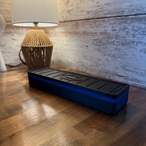 May include: A black rectangular box with a blue stripe, possibly a charging station or organizer, sits on a wooden surface. The box has a textured top with a design. The background includes a lamp with a beige shade and a wooden wall.