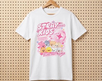 Può includere: Una t-shirt bianca a maniche corte con una stampa grafica rosa che riporta il testo STRAY KIDS, caratteri coreani e un gruppo di otto colorati personaggi animali dei cartoni animati. Il design include stelle, bolle e testo in inglese e coreano.