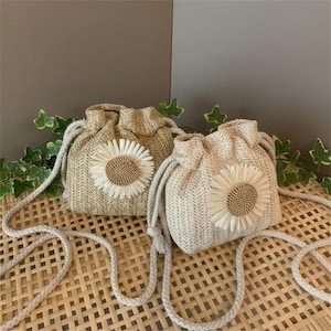 Puede incluir: Dos bolsas de tejido con cordón en beige y crema, cada una adornada con una aplicación de girasol. Cuentan con correas de hombro de cuerda. Los girasoles tienen centros marrón claro y pétalos blancos. Las bolsas están sobre una superficie tejida.