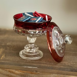 May include: A red and clear glass candy dish with a pedestal base and a matching removable lid. The bowl is filled with small, rectangular milk chocolate bars wrapped in blue and red paper, featuring a bear graphic and the text Milk Chocolate.