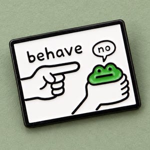 May include: A rectangular enamel pin with a white background and a black border. The design features a hand pointing at a green frog held in another hand. The text "behave" is printed in black, and a speech bubble above the frog contains the word "no".