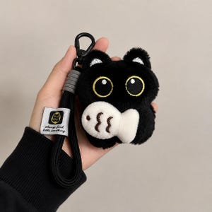 May include: A plush black cat-shaped case with large yellow eyes and a white fish-shaped accent. It features a black wrist strap with a grey connector and a black metal clip. A white fabric tag displays a cat icon and the text always find little something.