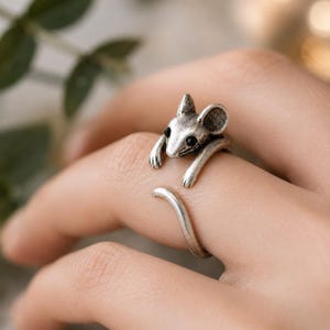 May include: A silver-toned ring featuring a detailed mouse design. The mouse has small black eyes and is wrapped around the finger, with its tail curving around the band. The ring is a unique piece of jewelry.