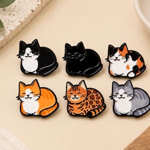 May include: A set of six enamel pins shaped like cats in various colors and patterns. The collection includes a black and white cat, a solid black cat, a calico cat, an orange tabby cat, a spotted orange cat, and a grey striped tabby cat.