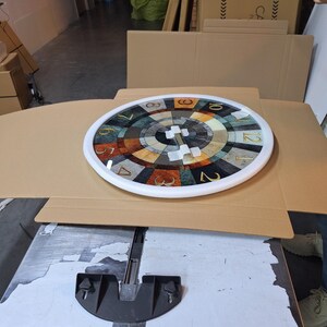 May include: A round wall clock with a white frame and a multi-colored, segmented face featuring a radial pattern of brown, gray, and cream tones. The clock face displays gold-colored Arabic numerals from one to twelve around the perimeter.