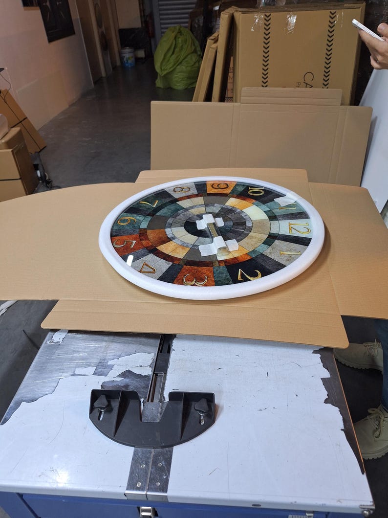 May include: A round wall clock with a white frame and a multi-colored, segmented face featuring a radial pattern of brown, gray, and cream tones. The clock face displays gold-colored Arabic numerals from one to twelve around the perimeter.