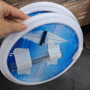 May include: A round wall clock featuring a blue graphic of a sailboat on the water. The clock face has white numbers and is protected by a white foam ring and clear plastic packaging secured with white adhesive tape for shipping purposes.