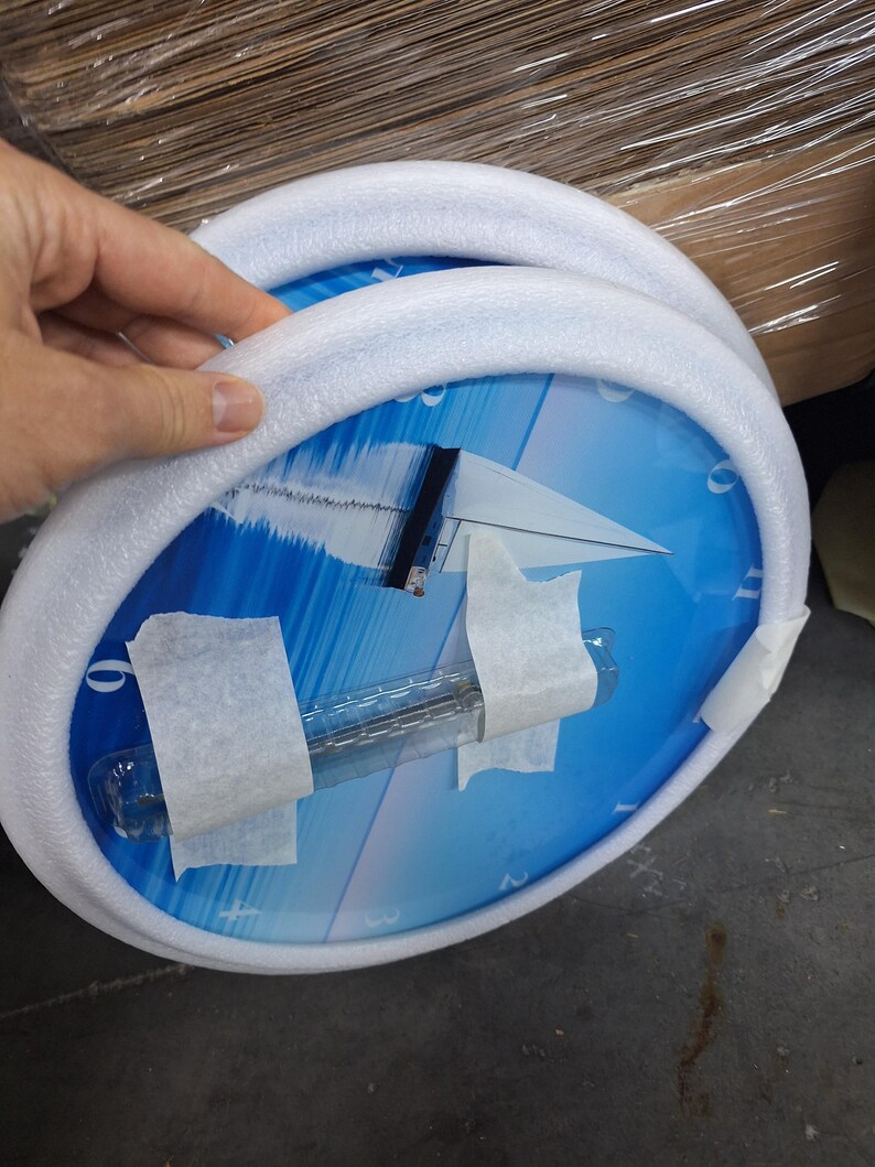 May include: A round wall clock featuring a blue graphic of a sailboat on the water. The clock face has white numbers and is protected by a white foam ring and clear plastic packaging secured with white adhesive tape for shipping purposes.