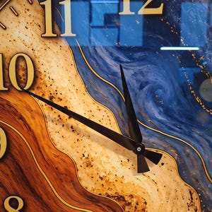 May include: A wall clock face featuring a swirling abstract pattern of deep blue, sandy beige, and dark brown tones. The clock has large gold-colored numerals for 8, 10, 11, and 12, with two thin black hands pointing toward the center.