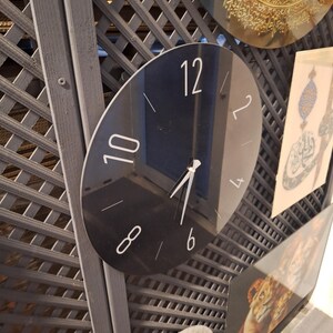 May include: A circular black wall clock with a reflective surface, featuring white Arabic numerals at the 12, 10, 8, 6, 4, and 2 positions, along with thin white hour and minute hands pointing toward the center of the clock face.