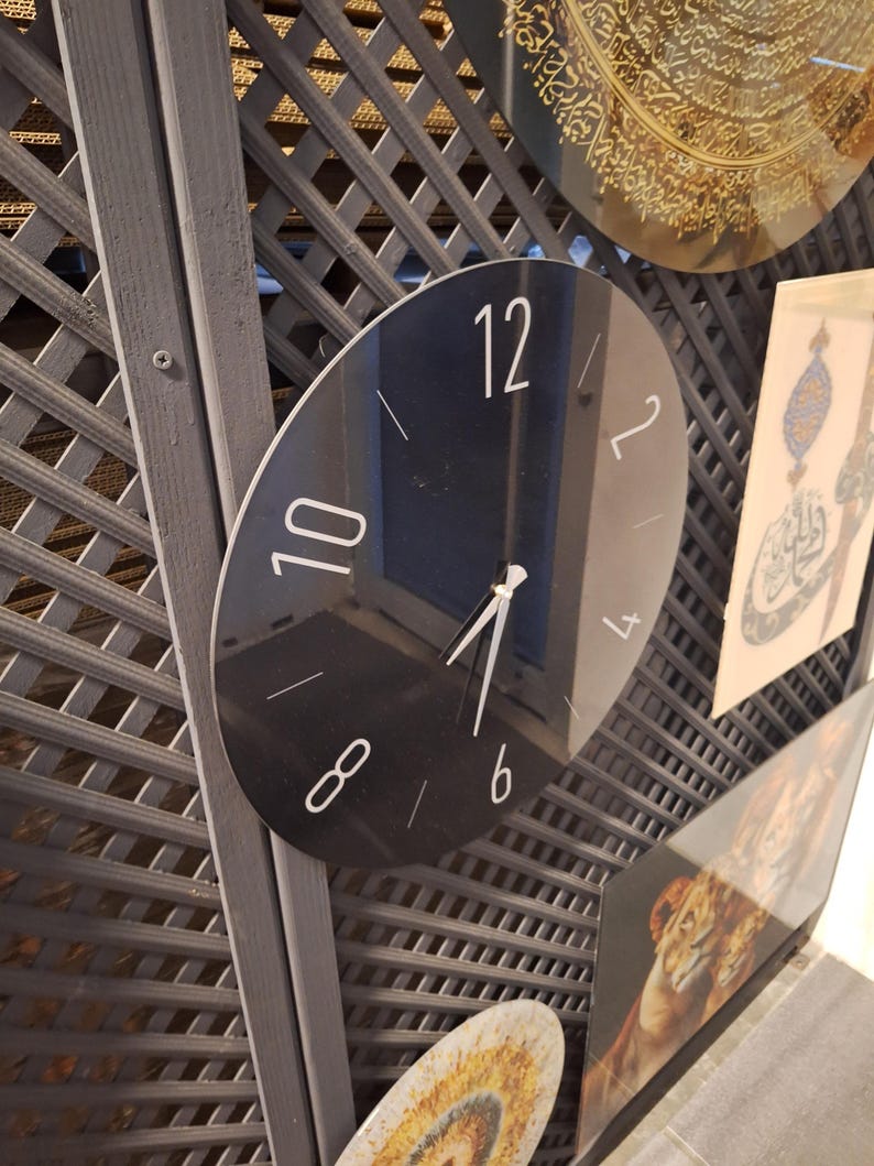 May include: A circular black wall clock with a reflective surface, featuring white Arabic numerals at the 12, 10, 8, 6, 4, and 2 positions, along with thin white hour and minute hands pointing toward the center of the clock face.
