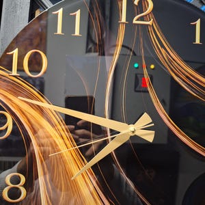 May include: A round wall clock with a black face featuring gold-colored numerals and a swirling orange and gold abstract pattern. The clock has gold-colored hour, minute, and second hands centered on the dial, with a reflective glass surface.