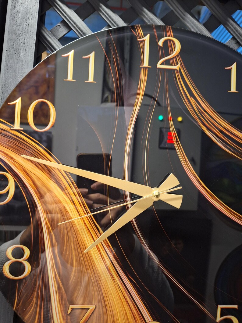 May include: A round wall clock with a black face featuring gold-colored numerals and a swirling orange and gold abstract pattern. The clock has gold-colored hour, minute, and second hands centered on the dial, with a reflective glass surface.