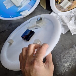 May include: A circular white foam clock base featuring a black battery-operated clock mechanism mounted in the center. Two small white foam spacers are attached to the surface, and white adhesive tape is applied to the back of the clock.