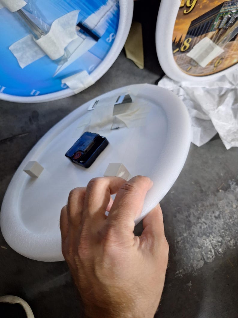 May include: A circular white foam clock base featuring a black battery-operated clock mechanism mounted in the center. Two small white foam spacers are attached to the surface, and white adhesive tape is applied to the back of the clock.