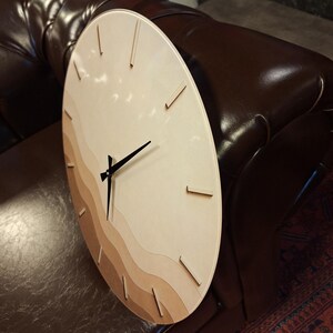May include: A circular wall clock featuring a light wood face with layered, wavy horizontal bands in varying shades of beige and tan. The clock has thin, dark brown hour markers and three slender black hands positioned at the center.