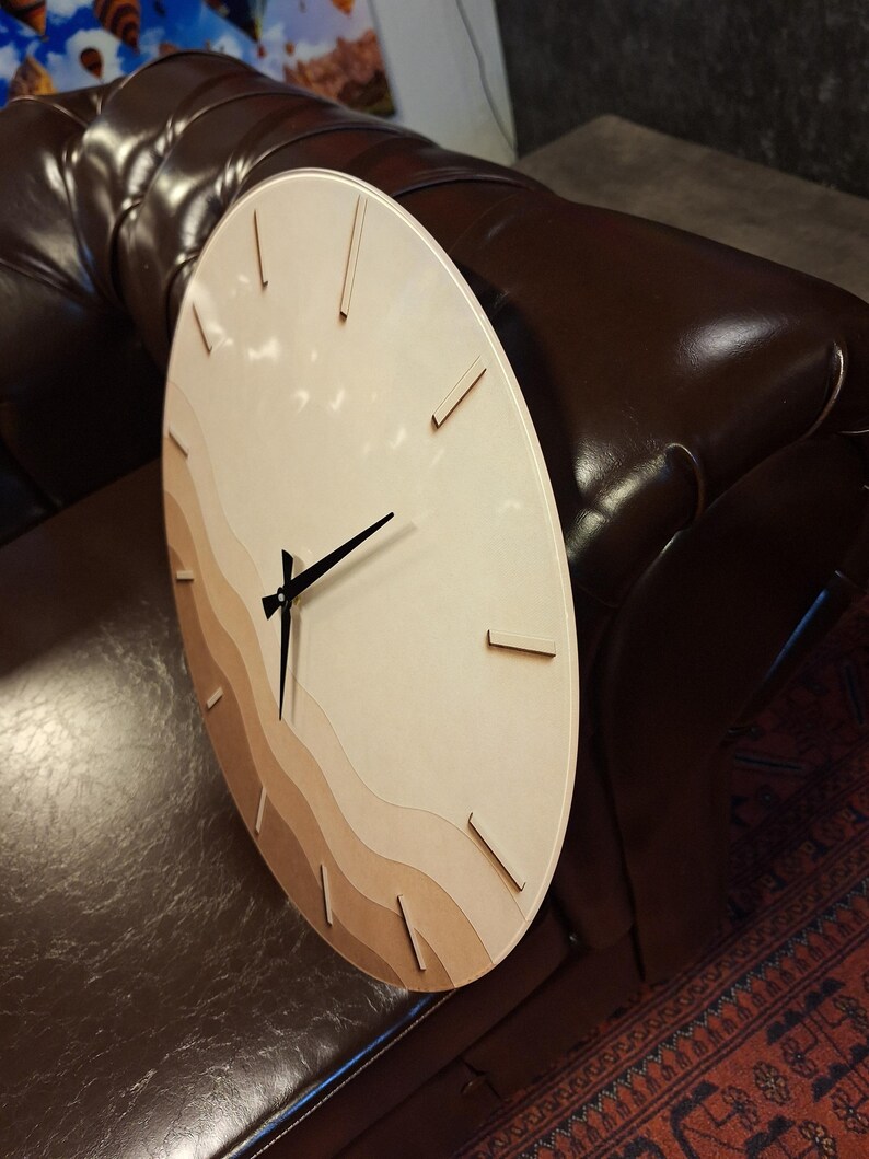 May include: A circular wall clock featuring a light wood face with layered, wavy horizontal bands in varying shades of beige and tan. The clock has thin, dark brown hour markers and three slender black hands positioned at the center.