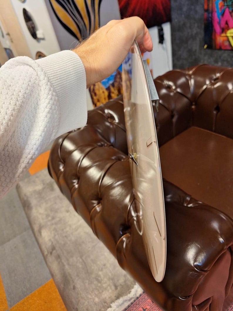 May include: A circular wall clock with a smooth, light-colored face and thin, dark hands. The clock has a slim profile and a metallic mechanism visible on the back, showing a side-view perspective of the timepiece's flat, disc-like structure.